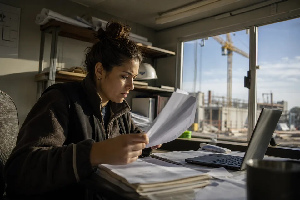 Construction company accountant in a job site trailer reviewing bank statements and check stubs beside a laptop