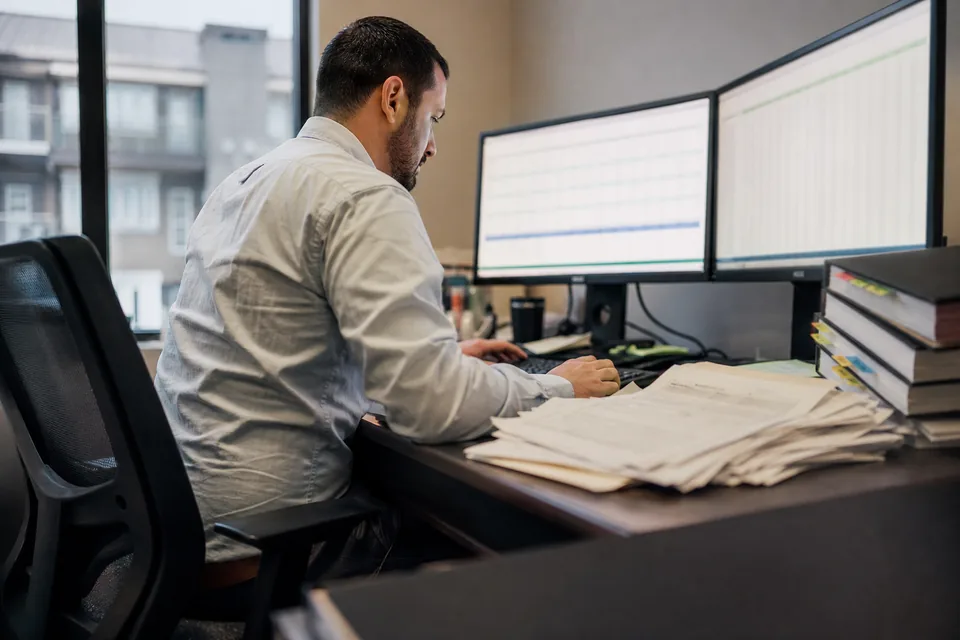 Property accountant reconciling bank statements for several rental properties at a desk with two monitors